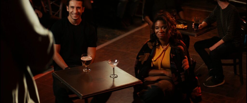 Akilah Hughes at comedy club sitting at a table looking at the stage
