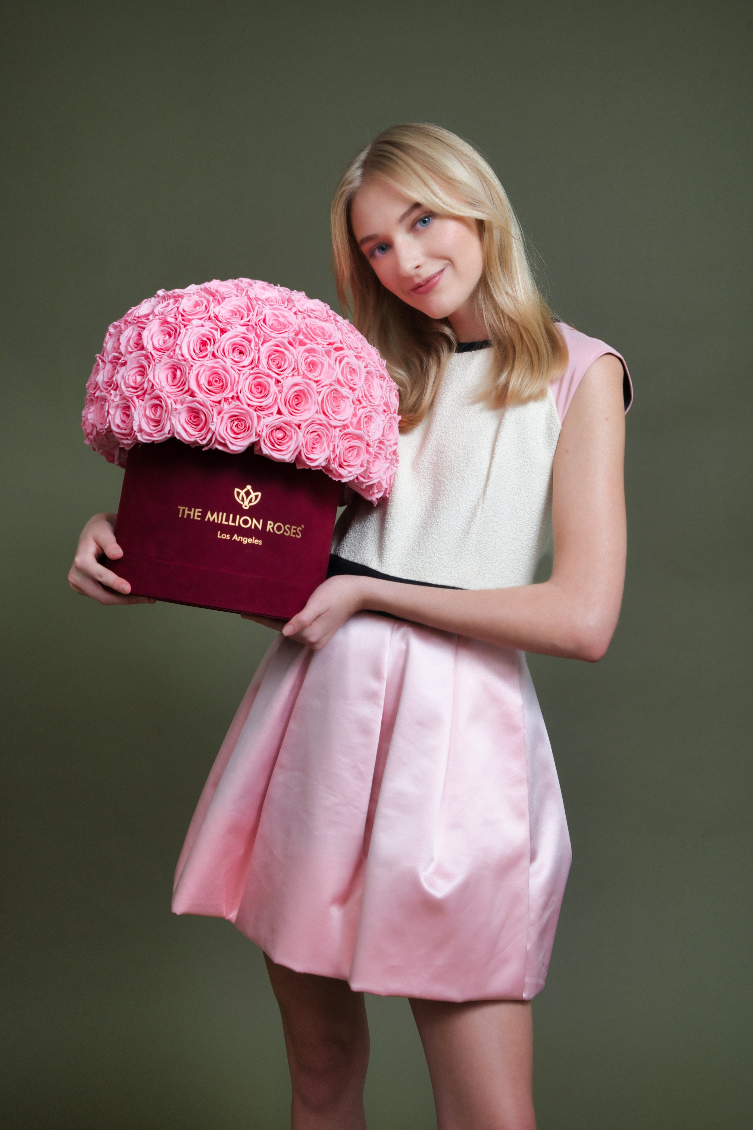 Photo model in white and pink dress presenting large pink The Million Roses flowers in red box