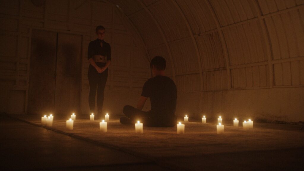 Person sitting on floor surrounded by candle lights facing a person standing upfront of him