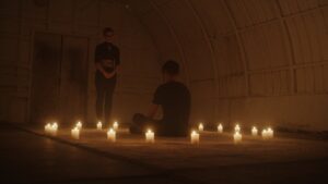 Person sitting on floor surrounded by candle lights facing a person standing upfront of him