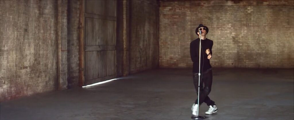 Wide shot of Sonny dancing in empty warehouse with microphone