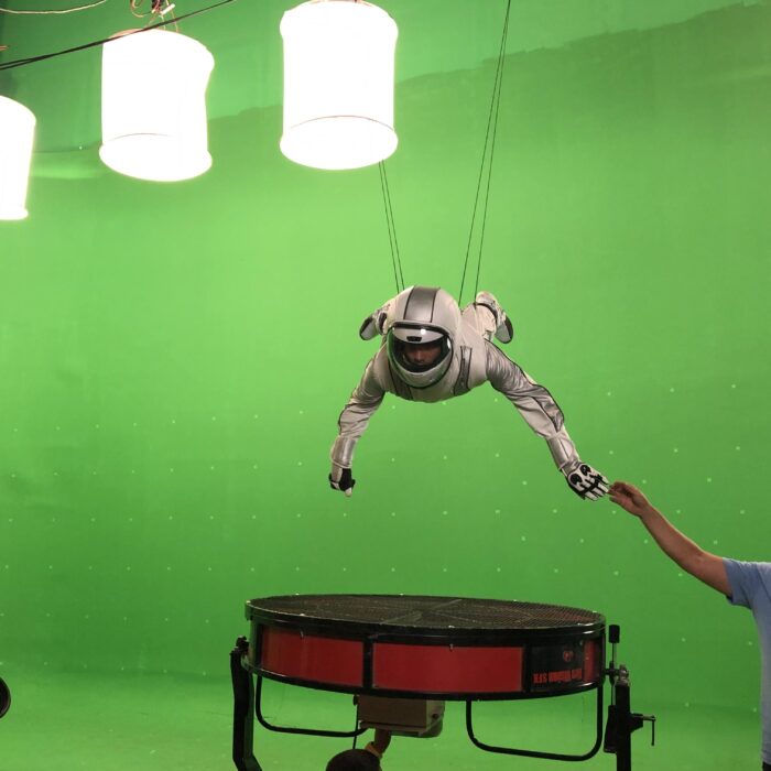 Greenscreen studio with stunt double suspended from wires. Fans below him. lights hanging from ceiling. stunt cordinator holding actor in space suite in place.